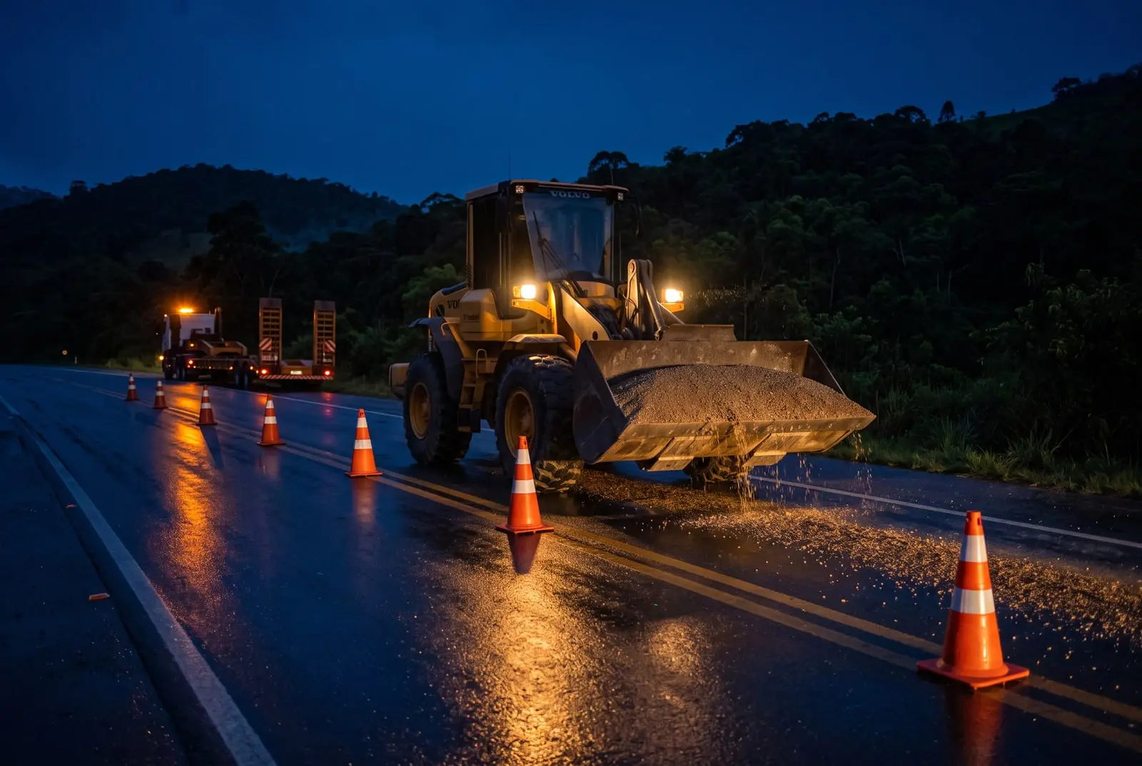 Pá carregadeira recolhendo o cascalho caído numa rodovia molhada à noite, com cones e uma prancha ao fundo